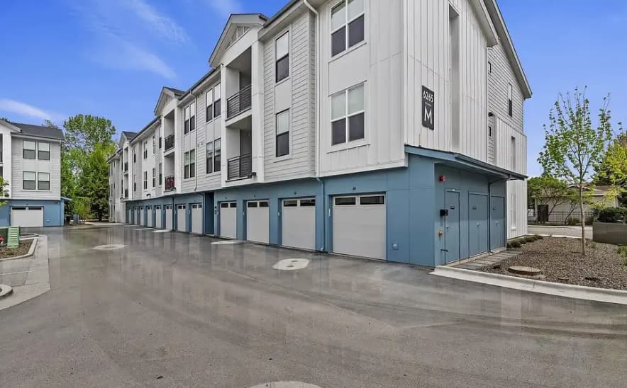 Modern three-story apartment building, River Pointe Apartments Boise, with white siding and blue trim, featuring multiple garages on the ground level and balconies above, set along a clean, paved street with trees and landscaping.