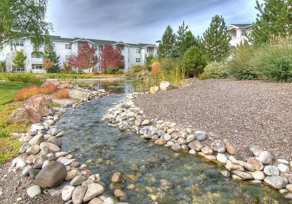A shallow stream with clear water and rocks winds through a landscaped garden at River Quarry Apartments Boise, surrounded by trees and shrubs. In the background, white apartment buildings stand under a cloudy sky.