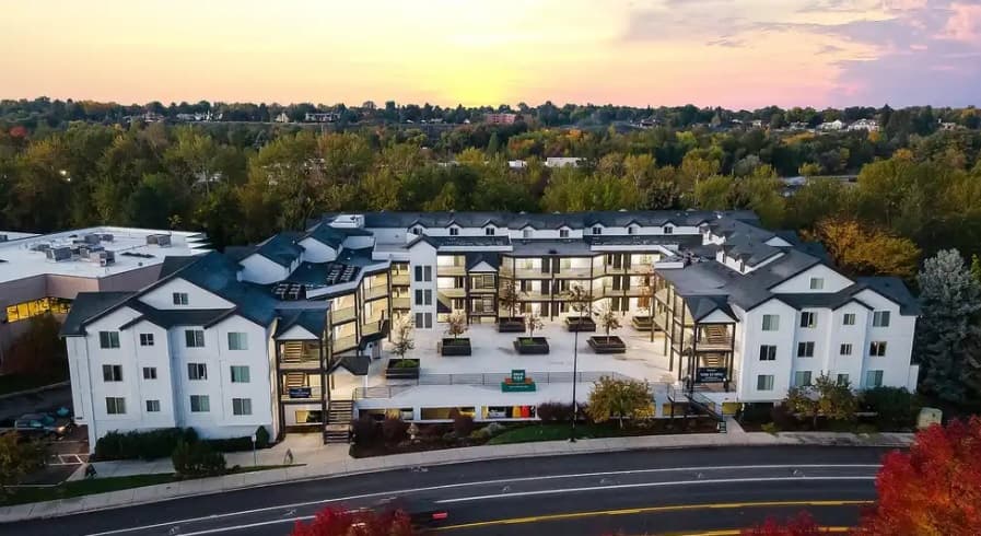 Aerial view of River Walk Apartments Boise, a large, modern complex with white exterior walls and dark roofs, nestled among trees and greenery at sunset, with a road in front.