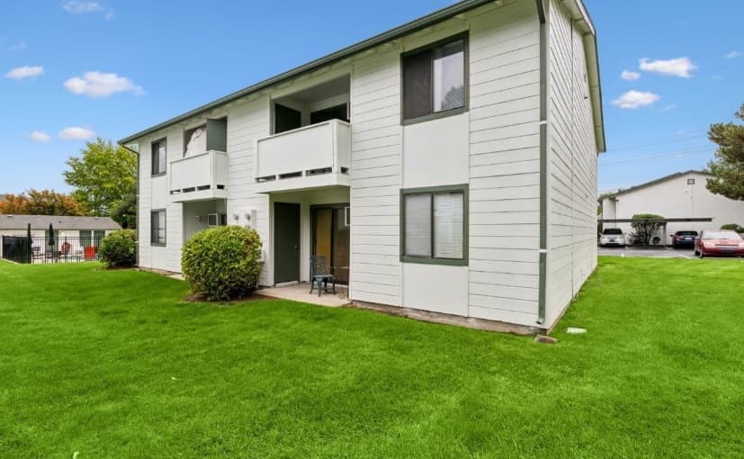 A two-story light gray apartment building with green trim, small balconies, and windows is nestled in Shannon South Boise, surrounded by a well-maintained grassy lawn under a partly cloudy sky. Cars and trees are visible in the background.