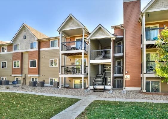 A modern, three-story apartment building at Village on Shields Fort Collins features tan and brown siding, multiple balconies, exterior staircases, and a grassy area with a sidewalk in the foreground.