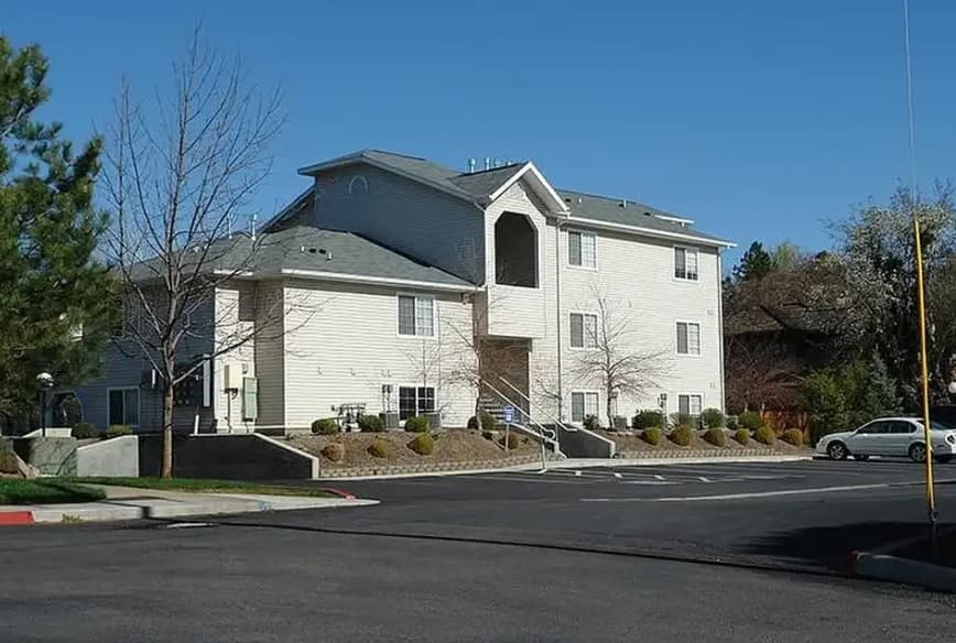 A two-story beige apartment building with a white staircase and railings, surrounded by shrubs and trees, sits on a clear day beneath a blue sky at Spring Creek Apartments Boise. A car is parked on the right side of the paved driveway.