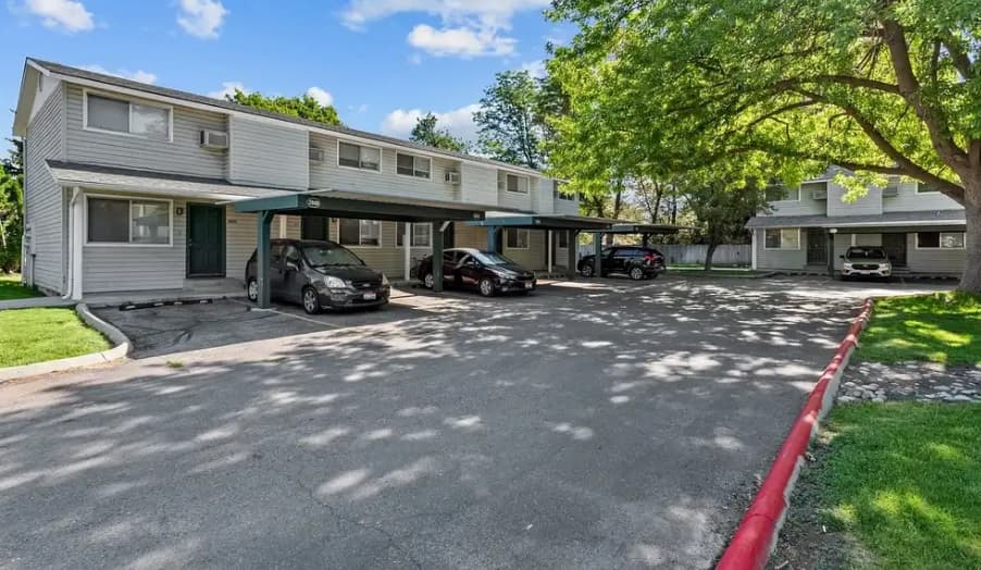 Sugar Pine Boise apartment complex features two-story buildings, covered parking spaces, several cars parked beneath large shade trees, and a clear blue sky with scattered clouds.