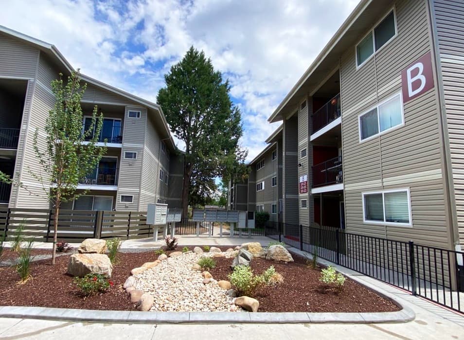 A landscaped courtyard with rocks and small plants sits between two beige, three-story Talavera Apartments Boise buildings labeled with the letter B on the right. The sky is partly cloudy.