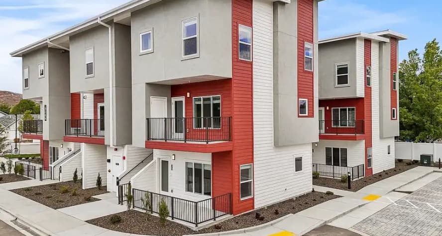 Modern three-story townhouses at The Edge at State feature red and white siding, large windows, balconies, and stairways leading to the entrances. These Apartments & Townhomes Boise offer minimal landscaping and a convenient parking area.