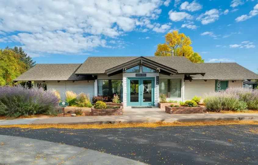 A single-story building with light siding, large front windows, a teal door, and landscaped flowerbeds sits under a blue sky with scattered clouds and autumn trees—the perfect welcome to The Hewing Fort Collins.
