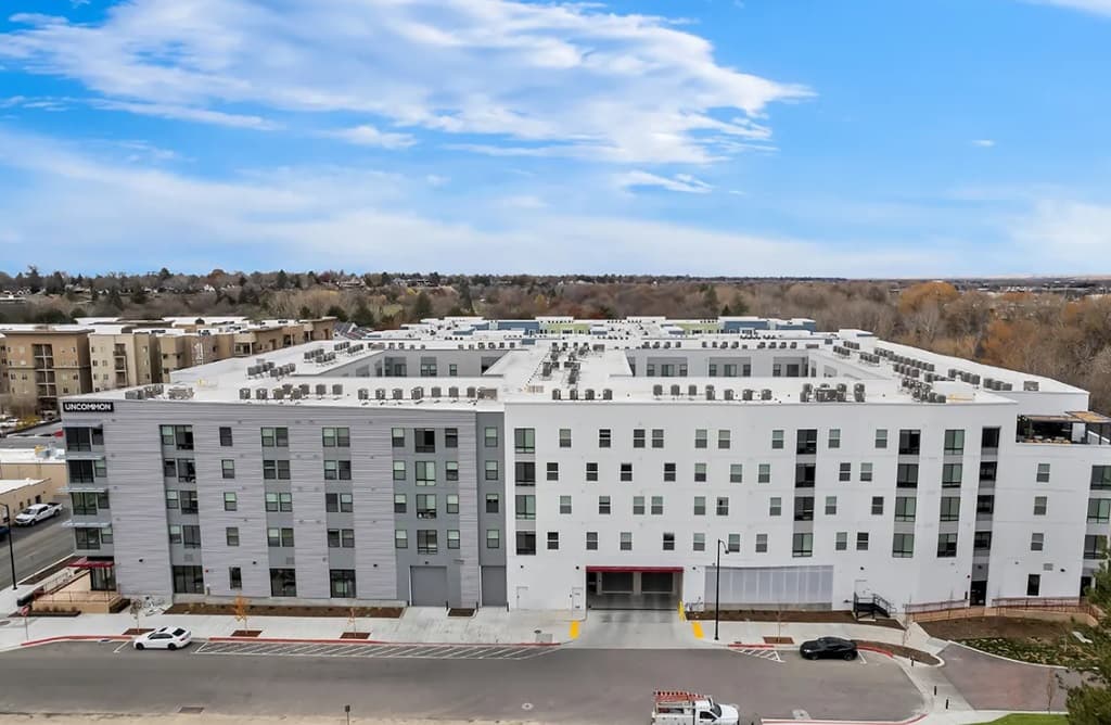 A modern, multi-story apartment building with a white and gray facade, large windows, and rooftop HVAC units sits next to a street in UNCOMMON BOISE, surrounded by trees under a partly cloudy sky.