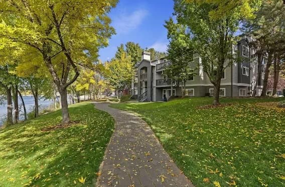 A paved path winds through a grassy area with scattered yellow leaves, flanked by tall trees with green and yellow foliage, leading to Whitewater Park Apartments Boise near the river under a blue sky.