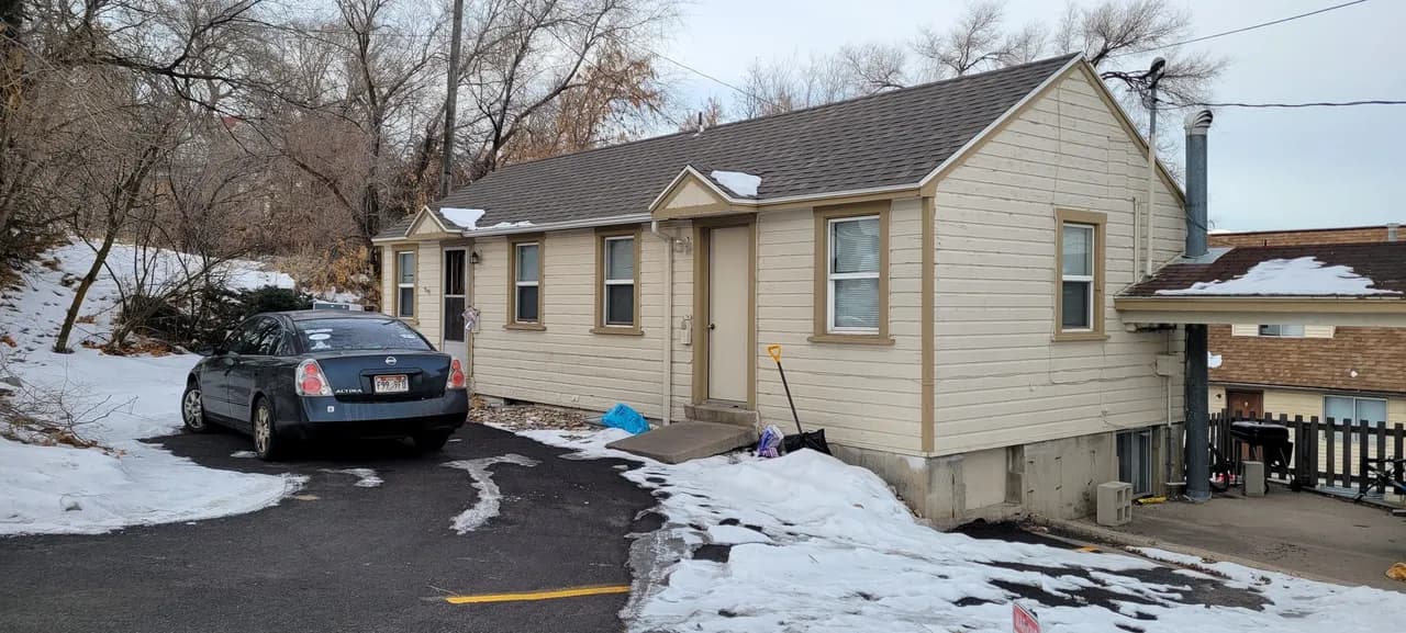 A small beige house with a shingled roof sits next to a snowy driveway at Auburn Heights Studio Logan. A black car is parked outside, snow blankets the ground, leafless trees surround the area, and small items are scattered near the house.