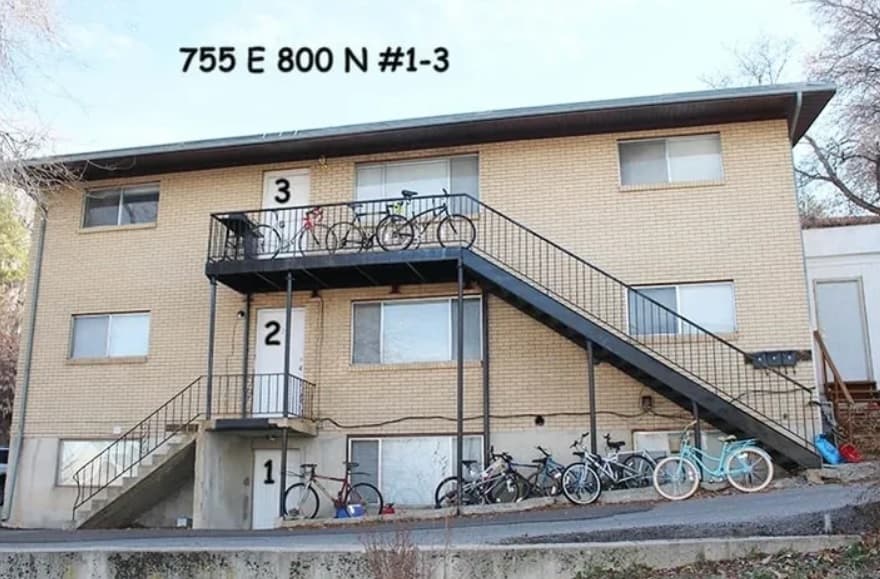 A two-story brick apartment building at Auburn Heights Terrace Logan with a black staircase leading up. Several bicycles are parked along the ground and on the balcony. The text 755 E 800 N #1-3 is at the top of the image.