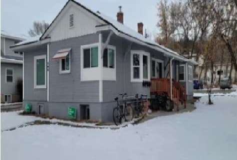 A small gray house with white trim sits on a snowy lawn in Autumn Hills by USU Logan. Two bicycles are parked next to the porch, which has wooden stairs leading to the front door. Trees and neighboring houses are visible in the background.
