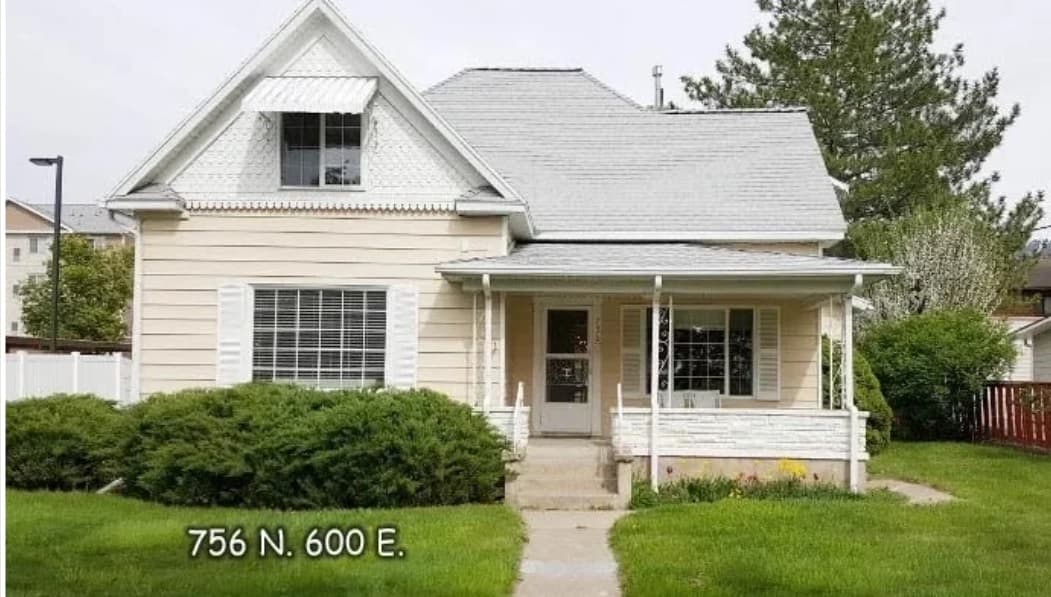 A beige, two-story Austin Ridge Doll House sits behind green bushes and a well-kept lawn in Logan, with white trim and a covered front porch. The address 756 N. 600 E. appears in white text on the lower left.