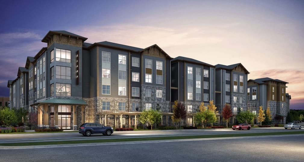 Modern four-story apartment building at dusk, with large windows, stone and wood exterior, landscaped trees and shrubs, and cars parked along the street—welcome to Elara at The Sawmill Flagstaff.