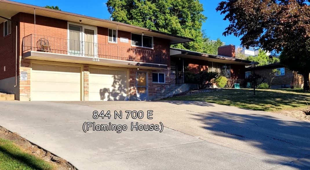A two-story brick house at Austin Ridge Flamingo, featuring two garage doors, a balcony, and sloped driveway. Text on the image reads 844 N 700 E (Flamingo House). Trees and lawn surround this Logan home.