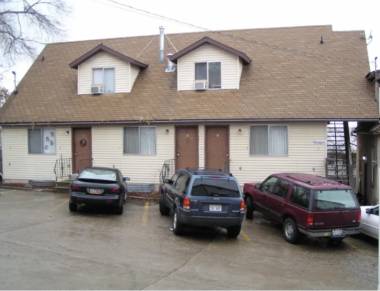 Three parked cars sit in front of a beige two-story duplex at Auburn Heights, 782 Logan, with brown trim and matching front doors. The wet driveway and parking area hint at recent rain on this cloudy day.