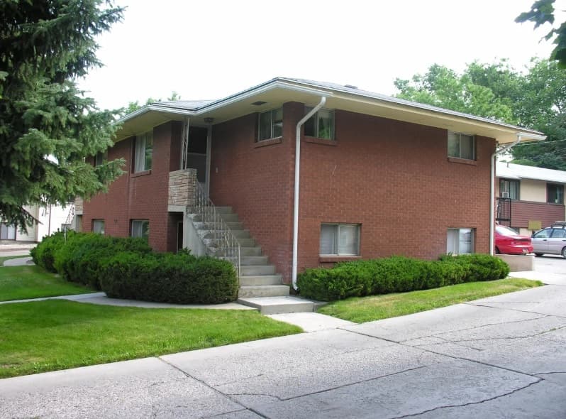 The Austin Ridge Lanny 2 Bed is a two-story brick apartment building with external stairs, neatly trimmed bushes, and green lawns, located beside a paved parking area with parked cars.