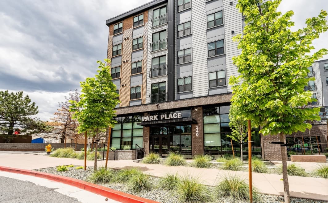 A modern mid-rise apartment building named Park Place Flagstaff with large windows, young trees, and landscaped plants in front. The street number 240 is visible near the entrance. The sky is cloudy.