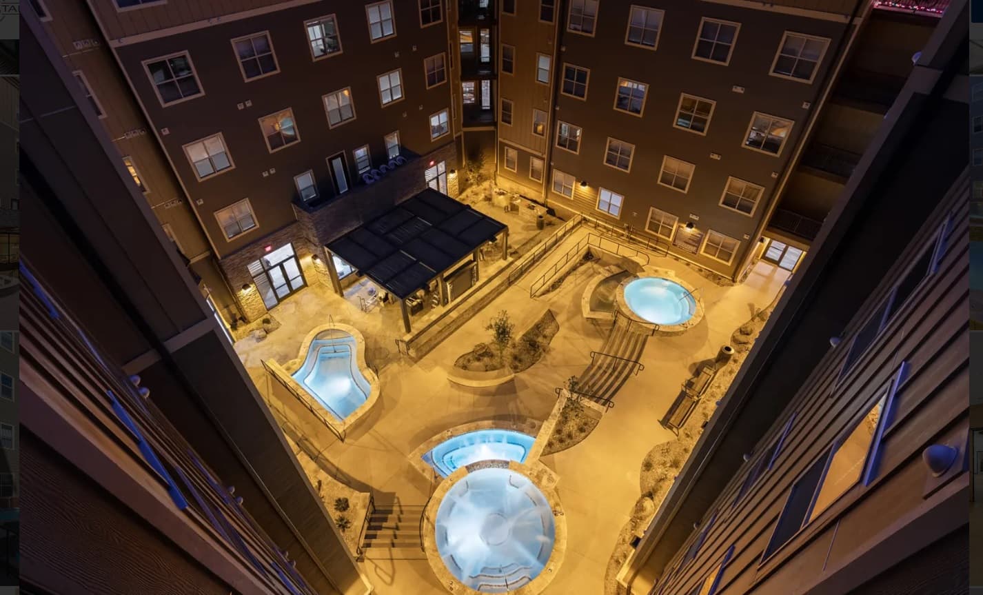 Night view of The Standard at Flagstaff courtyard with three illuminated hot tubs, seating areas, landscaping, and surrounding multi-story brick buildings with numerous windows in vibrant Flagstaff.