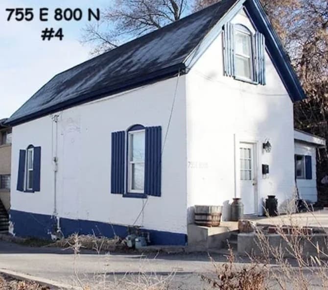 A small white Auburn Heights Terrace House with blue shutters and trim, a steep roof, and an upper window. The address 755 E 800 N #4 is written in black at the top left. Trees and dry grass fill the foreground in Logan.