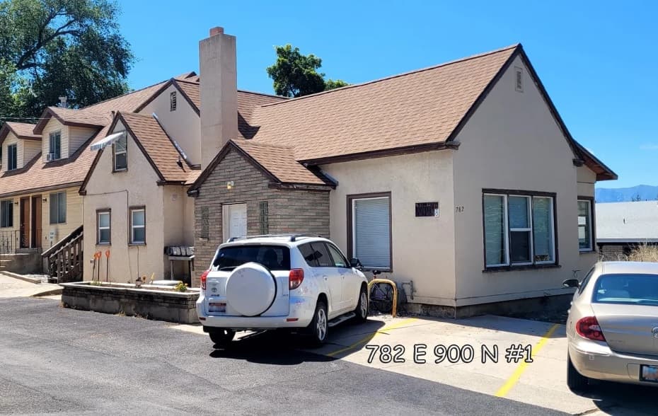 A beige Auburn Heights Track house with a brown roof and chimney, two cars parked in front, and 782 E 900 N #1 written on the image. Other houses and trees are visible in the background of Logan.