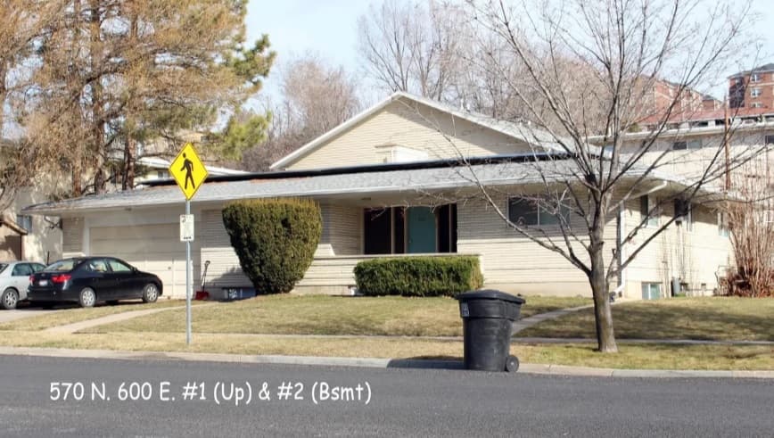 A single-story beige house with a porch, trimmed bush, and leafless trees sits near a pedestrian crossing sign in Logan’s Autumn Hills. Parked cars and a trash bin line the curb. Text reads: 570 N. 600 E. #1 (Up) & #2 (Bsmt).