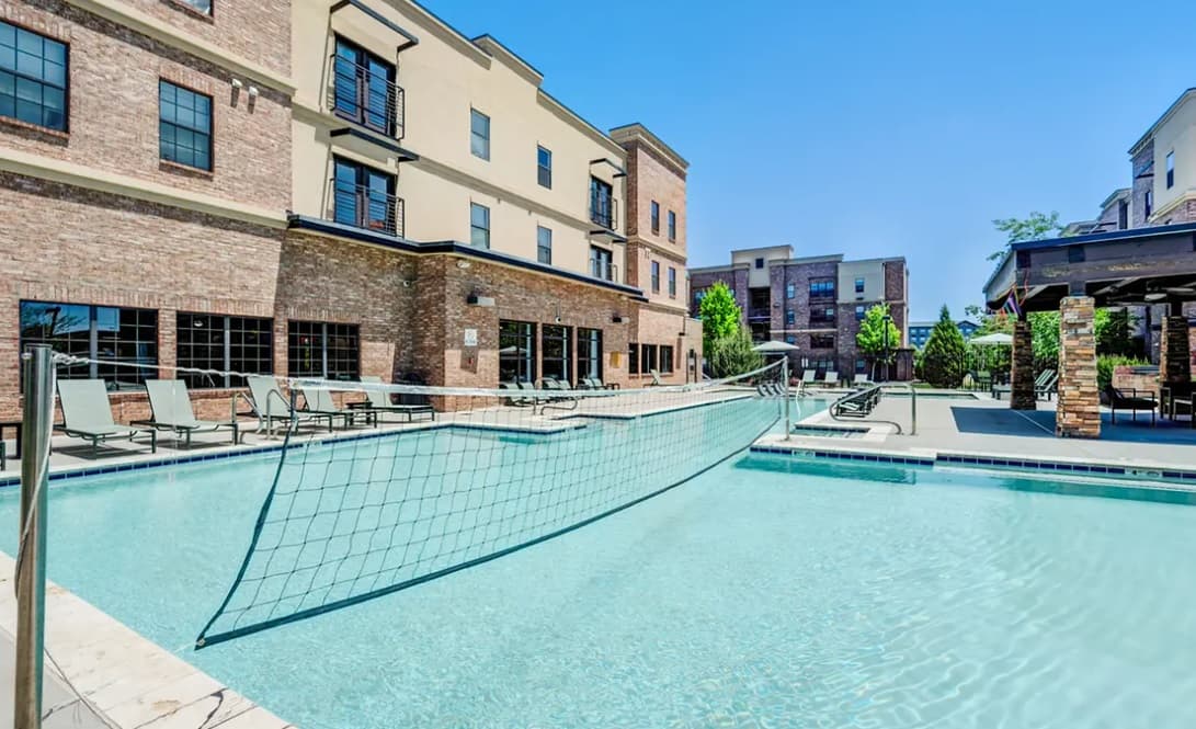 A swimming pool with a volleyball net stretched across it sits at Yugo Flagstaff Grove, surrounded by lounge chairs and modern apartment buildings under a clear blue sky.