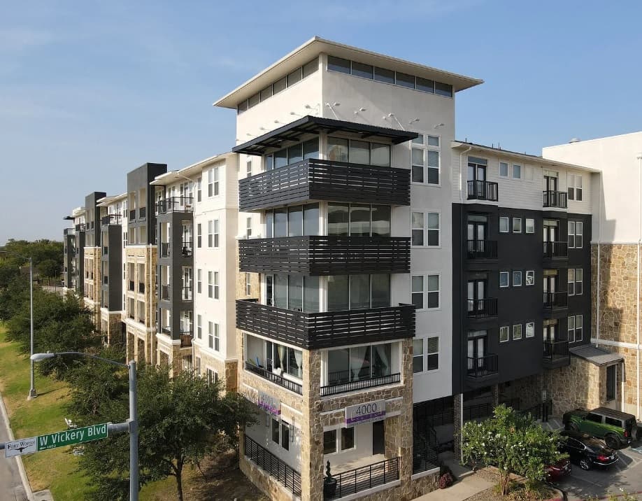 A modern four-story apartment building at 4000 Hulen Fort Worth features large balconies and a mix of white, black, and stone exterior. Located on W Vickery Blvd, trees line the street in front on a sunny day.