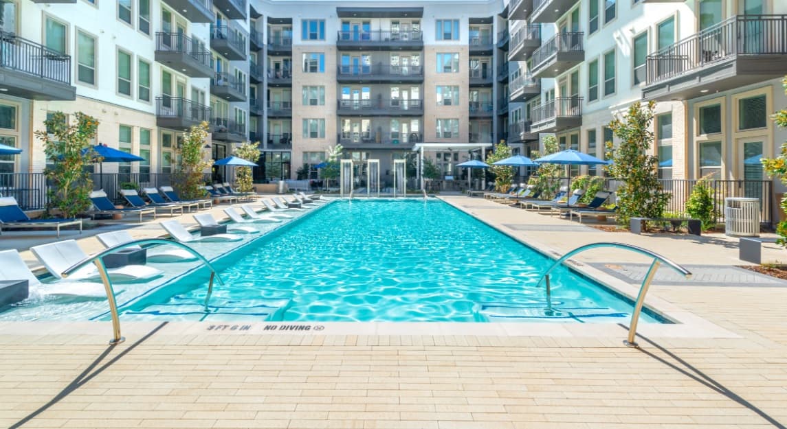 Modern apartment complex courtyard at Burnett Lofts Fort Worth featuring a large rectangular swimming pool with lounge chairs in the water, poolside seating, blue umbrellas, and surrounding multi-story buildings with balconies.