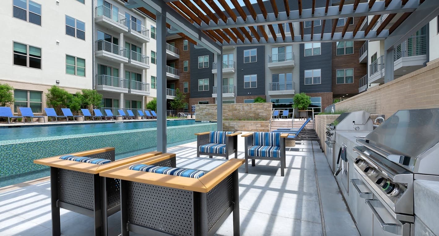 Modern outdoor lounge area at Atlas Waterside Fort Worth features striped cushioned chairs, stainless steel grills, and a pergola beside a swimming pool lined with blue lounge chairs, all surrounded by contemporary apartment buildings.