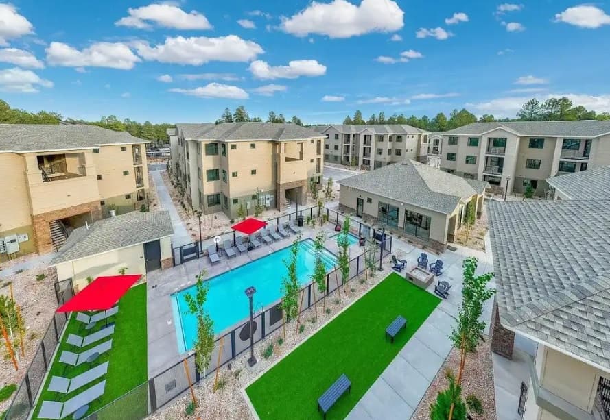 Aerial view of Aura Flagstaff, a modern apartment complex in Flagstaff with a central outdoor pool, lounge chairs, red umbrellas, green artificial turf, pathways, and three-story beige buildings surrounded by trees under a blue sky.