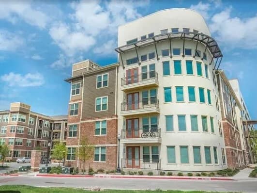 Modern apartment building at Aviator West 7th Fort Worth with multiple floors, large windows, and balconies, featuring a mix of brick and white exterior under a blue sky with scattered clouds.