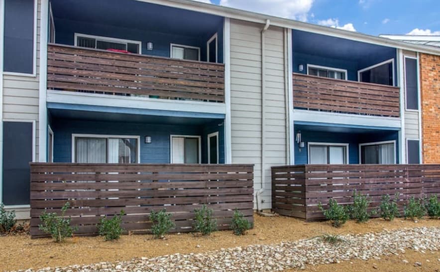 Two-story apartment building at The Barcelona On Chisholm Trail Fort Worth with dark wooden fenced patios and balconies, blue and white exterior walls, gravel ground cover, and small bushes in front.