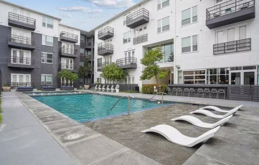Modern apartment complex courtyard at Bell Lancaster Apartments Fort Worth featuring a rectangular swimming pool, submerged lounge chairs, poolside seating, trees, and adjacent bar area with stools, surrounded by multi-story white and gray buildings.