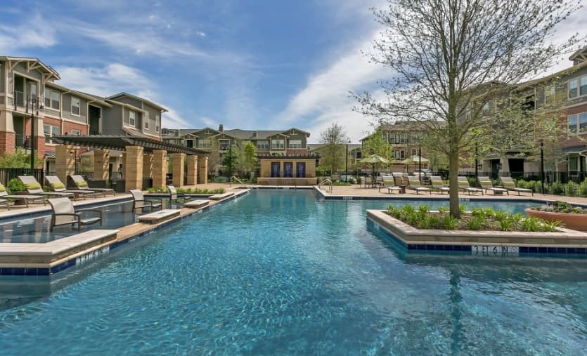 Outdoor swimming pool surrounded by lounge chairs, trees, and landscaped areas in the courtyard of The Berkeley Apartments Fort Worth under a blue sky with scattered clouds.