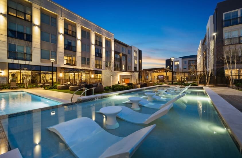 Modern apartment complex at dusk with illuminated buildings at Bexley Clearfork Fort Worth, featuring a long outdoor pool with white lounge chairs partially submerged in the water and landscaped greenery surrounding the area.