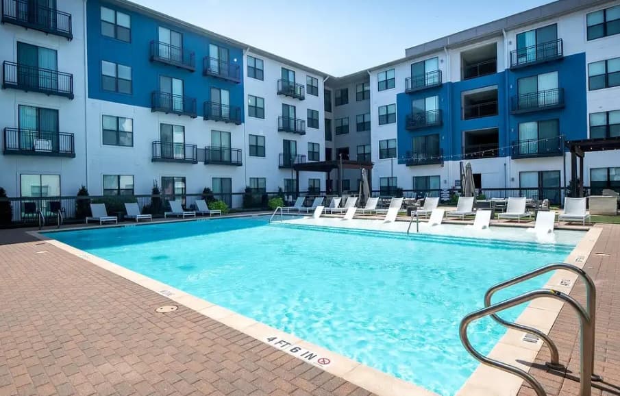 Broadstone on Trinity Fort Worth offers a modern apartment complex with a blue and white exterior, surrounding a large outdoor swimming pool with lounge chairs and umbrellas on the pool deck.