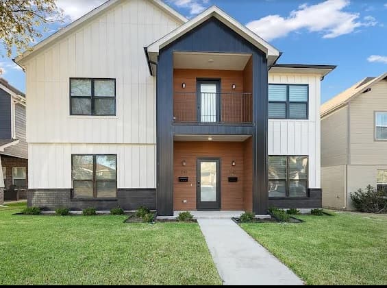 Modern two-story Casa Rana Fort Worth house with light siding, black trim, and wood accents. Features a covered entryway, small balcony above, black-framed windows, and a neatly manicured front lawn with a concrete walkway.