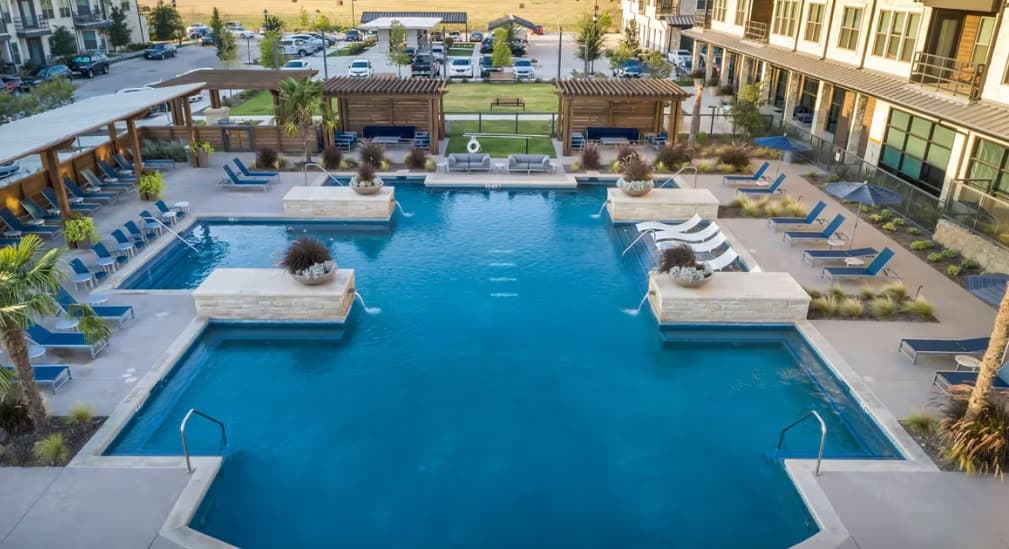 A large, modern outdoor swimming pool with lounge chairs, cabanas, and landscaped planters at Chisholm at Tavolo Park Fort Worth, surrounded by apartment buildings and a parking lot in the background.