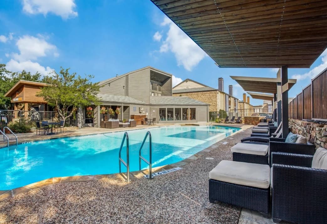 Outdoor swimming pool with clear blue water, surrounded by lounge chairs and shaded seating areas. Modern Chisholm Ranch Apartments Fort Worth buildings and trees are visible in the background under a partly cloudy sky.