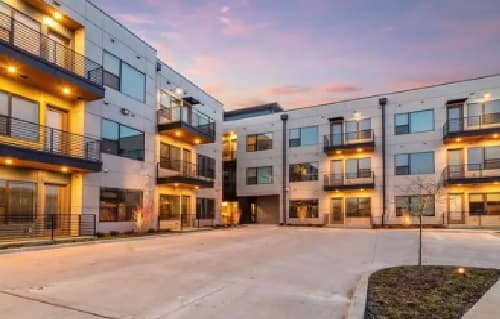 Modern Cityscape Apartment complex in Fort Worth with three-story buildings, large windows, and balconies glowing at dusk. The spacious courtyard features concrete pavement and minimal landscaping for a contemporary urban feel.