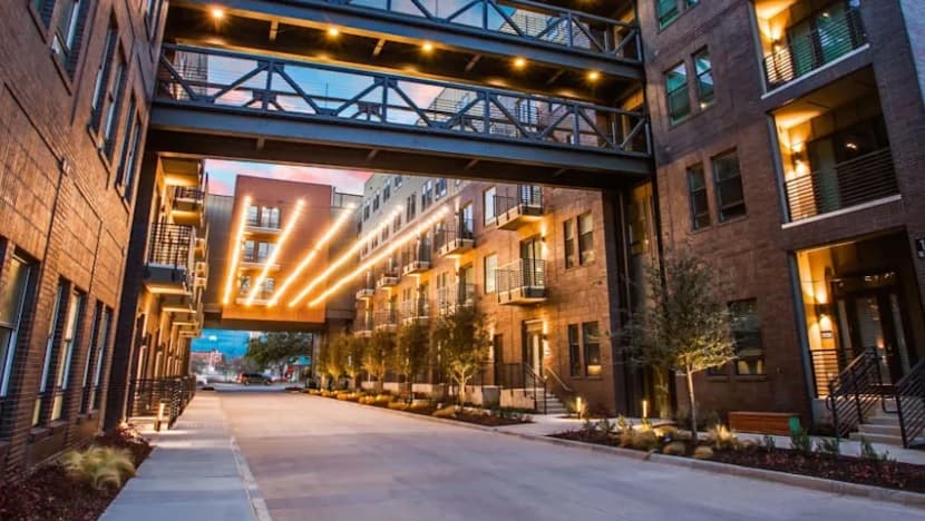 Modern apartment buildings with balconies line both sides of a clean, landscaped street at sunset near The Cooper Fort Worth. Two glass and metal skywalks connect the buildings above, with warm lights illuminating the scene.