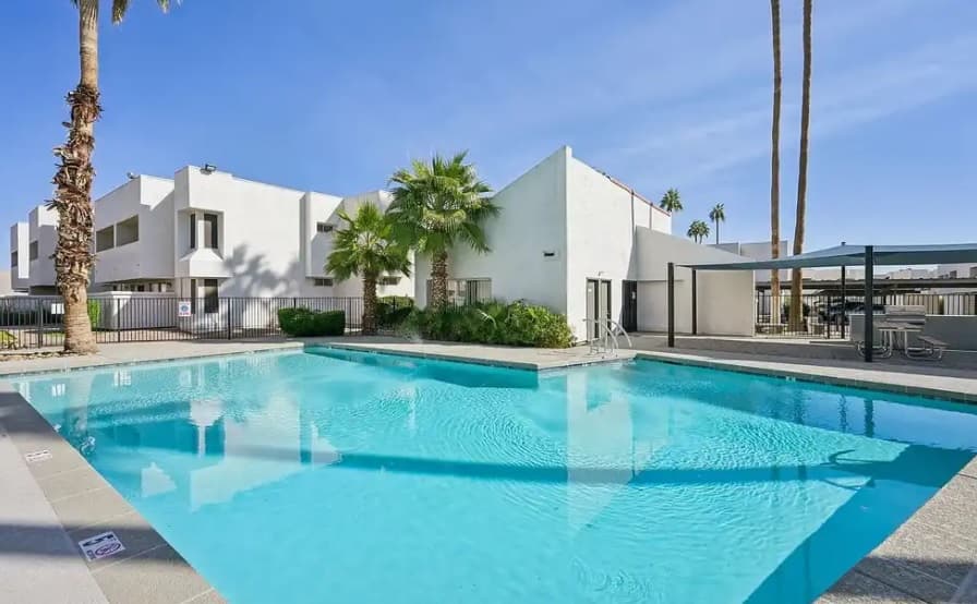 A clear blue outdoor swimming pool at Cove on 44th Apartments Phoenix is surrounded by palm trees and modern white buildings under a sunny sky, with shaded seating and a fence in the background.