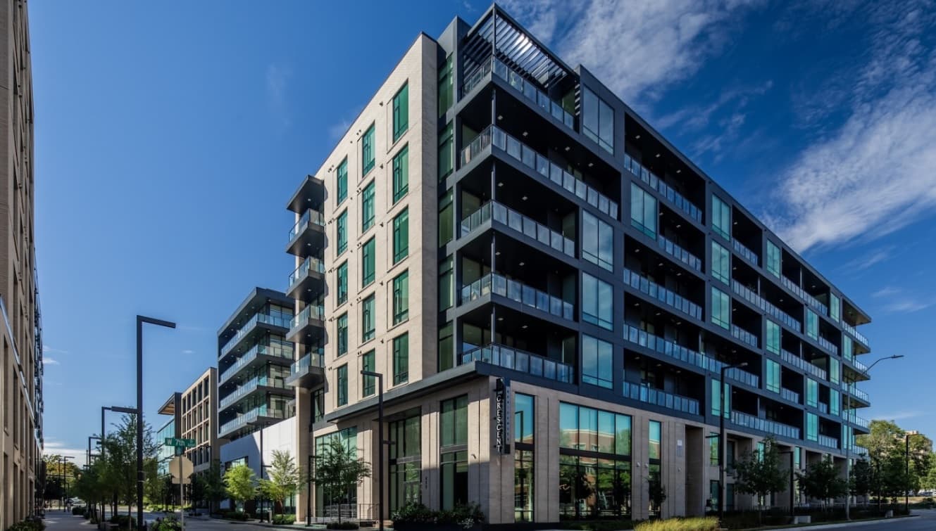 Modern mid-rise apartment buildings at The Crescent Residences Fort Worth feature large glass windows and balconies under a clear blue sky. Trees and streetlights line the sidewalk, with sunlight casting shadows on the contemporary architecture.