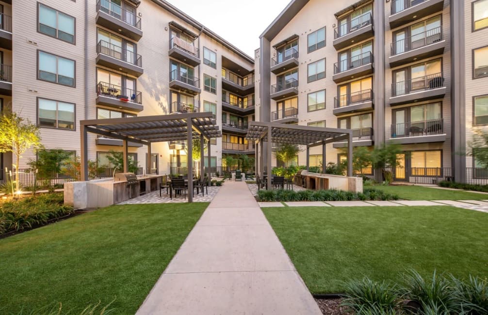 A landscaped courtyard at Crystal Springs Fort Worth features a pergola with seating areas, green lawns, lush plants, and surrounding multi-story buildings with balconies and large windows.