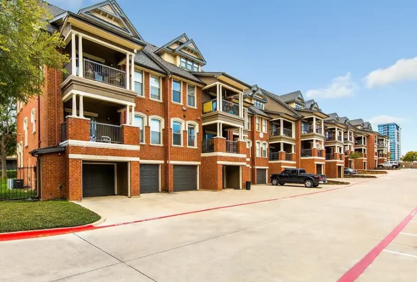 A row of modern, three-story red brick apartment buildings with balconies and attached garages lines the wide concrete driveway at Firestone at West 7th Fort Worth on a clear, sunny day.
