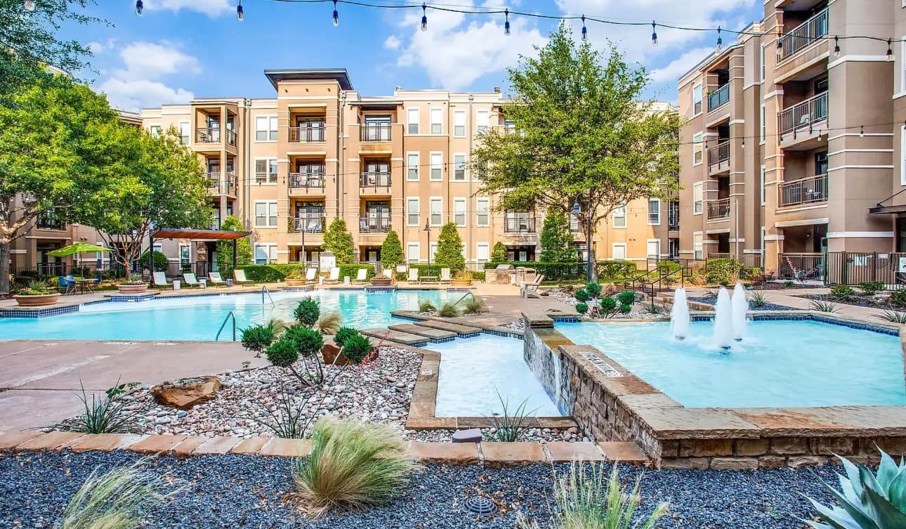 The Franklin at Samuels Avenue Fort Worth boasts a landscaped courtyard with a large swimming pool, a smaller fountain pool, lounge chairs, string lights above, and four-story buildings under a blue sky with scattered clouds.