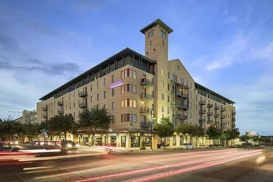 A multi-story apartment building, Grandmarc at Westberry Place Fort Worth, features retail shops on the ground floor, balconies on each level, a tall tower feature, and cars creating light trails on the street at dusk.