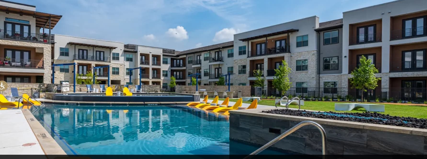 Modern apartment complex with three-story buildings surrounds a swimming pool at Hangar 19 Fort Worth. Yellow lounge chairs are partially submerged in the pool, with green lawns and a blue sky dotted with scattered clouds.