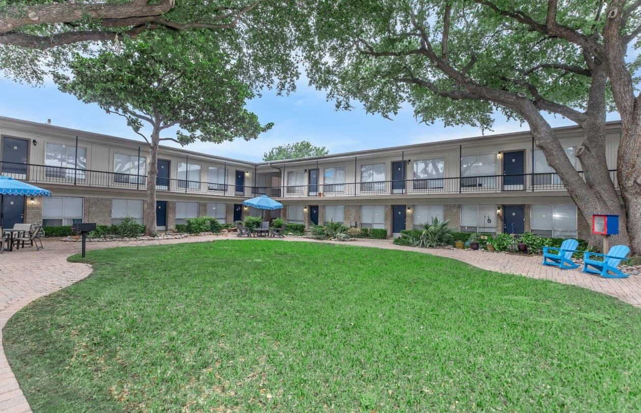 A grassy courtyard at Harris Gardens Fort Worth is surrounded by a two-story apartment building with outdoor walkways. Blue patio chairs and umbrellas provide seating, while large trees offer shade beneath the clear, bright sky.