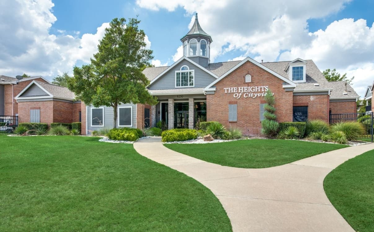 A brick building with a cupola and white trim, surrounded by green grass and landscaping. A curved sidewalk leads to the entrance, where Heights of Cityview Fort Worth is displayed on the wall.